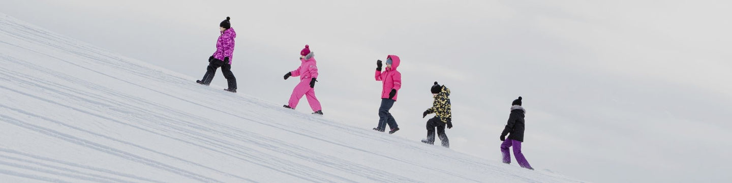 Viisi ihmistä värikkäissä talvivaatteissa kävelemässä lumisella rinteellä